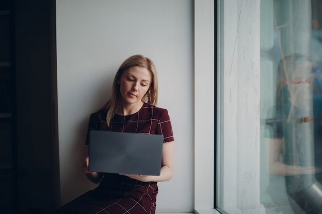 Jong mooi blond studentenmeisje met laptop zitting bij groot venster