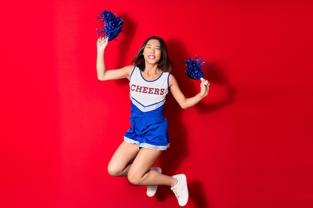 Jong mooi Chinees meisje glimlacht gelukkig in cheerleader uniform springt met een glimlach op het gezicht met pompon op een geïsoleerde rode achtergrond