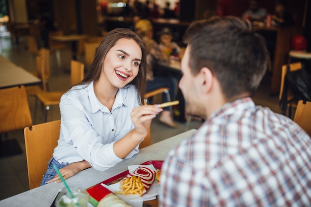 Jong, mooi donkerbruin meisje, glimlachend, voedt een jongen frietjes in een fastfoodcafé