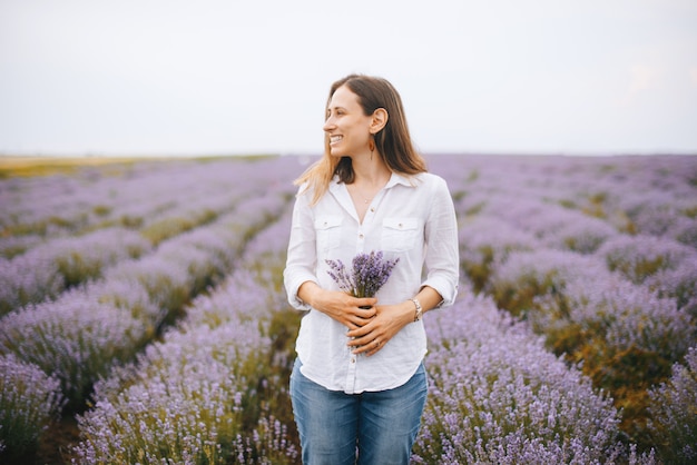 Jong mooi glimlachend meisje dat sommige lavendelbloemen houdt die ver weg kijkend zich, op een gebied van lavendel bevinden.