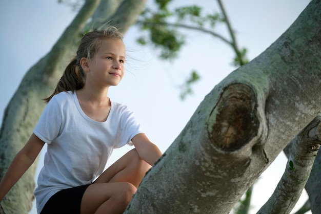 Jong mooi kindmeisje zit ontspannen tussen grote takken van oude boom op zonnige zomerdag