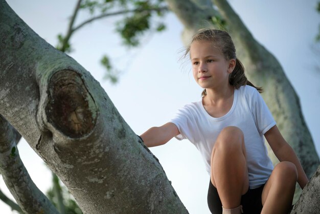 Jong mooi kindmeisje zit ontspannen tussen grote takken van oude boom op zonnige zomerdag