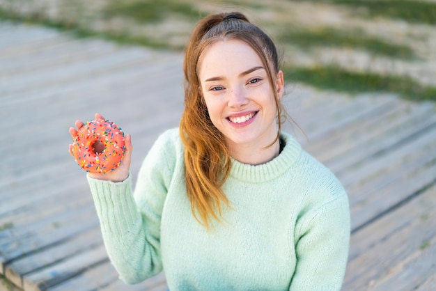 Foto jong mooi meisje dat een donut vasthoudt in de buitenlucht en veel glimlacht