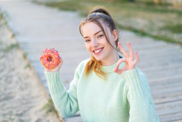 Jong mooi meisje dat een donut vasthoudt in de open lucht en ok teken met vingers laat zien