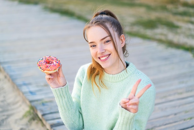 Foto jong mooi meisje dat een donut vasthoudt in de open lucht glimlachend en overwinningsteken toont