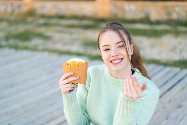 Jong mooi meisje dat gebakken frites buiten vasthoudt en uitnodigt om met de hand te komen blij dat je gekomen bent