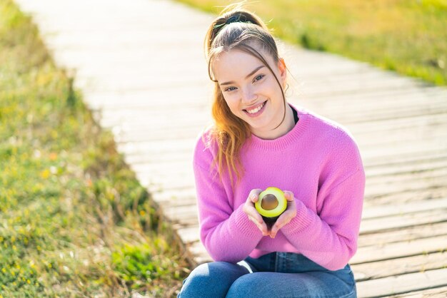 Jong mooi meisje dat in openlucht een avocado met gelukkige uitdrukking houdt