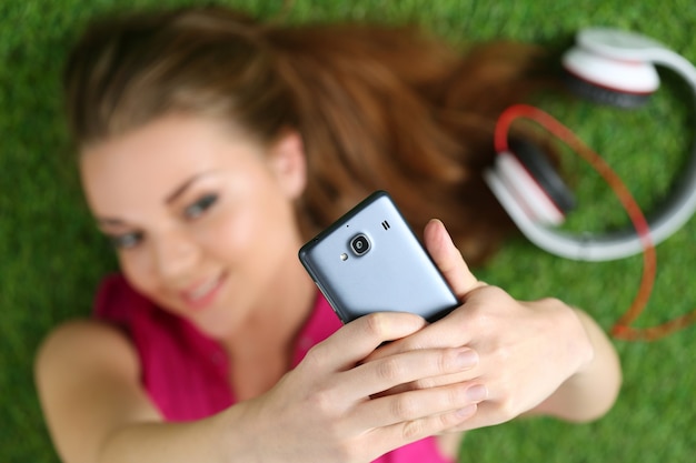 Foto jong mooi meisje dat zelfportret met haar slimme telefoon neemt die op gras legt. focus op telefoon