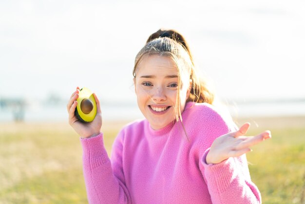 Jong mooi meisje houdt een avocado buiten met geschokte gezichtsuitdrukking