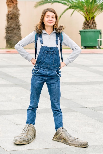 Foto jong mooi meisje in denim jumpsuit staat op de stoep in enorme schoenen