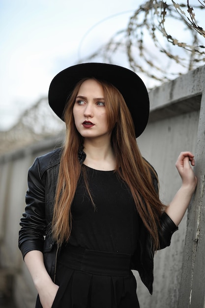 Jong mooi meisje in een hoed en met een donkere make-up buiten. Meisje in de gotische stijl op straat. Een meisje loopt door de stadsstraat in een leren vest met telefoon.