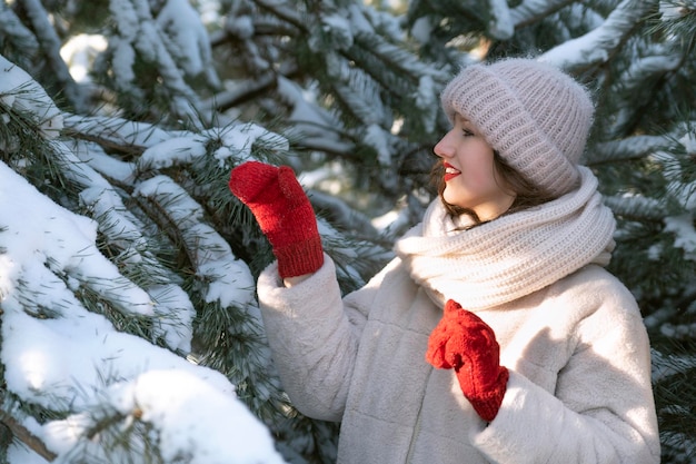 Jong mooi meisje in warme muts en rode wanten in de buurt van besneeuwde pijnbomen