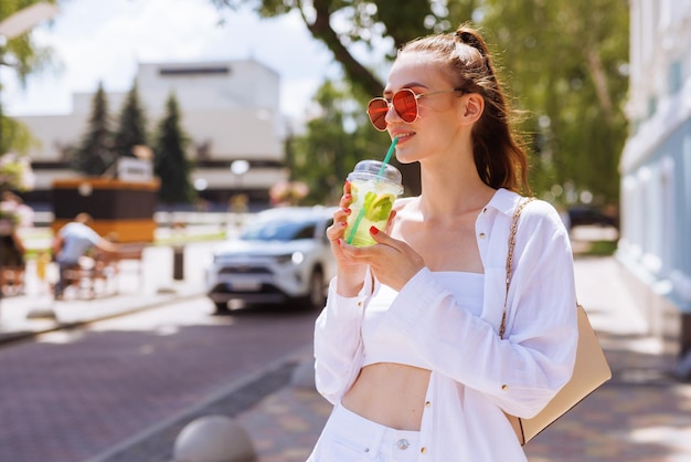 Jong mooi meisje in witte kleren en rode bril drinkt een zomerse verfrissende cocktail