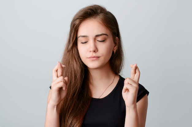 Jong mooi meisje in zwart shirt dragen gouden ketting en oorbellen kruising haar vingers en wensen voor geluk of dromen van iets