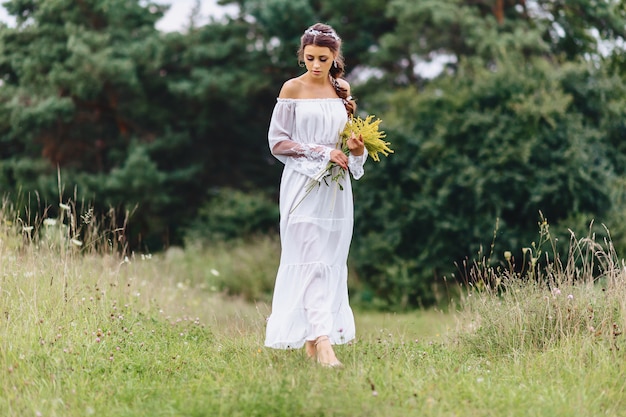 Jong mooi meisje met bloem in lichte het kleden gang bij gazon dichtbij de zomerbos