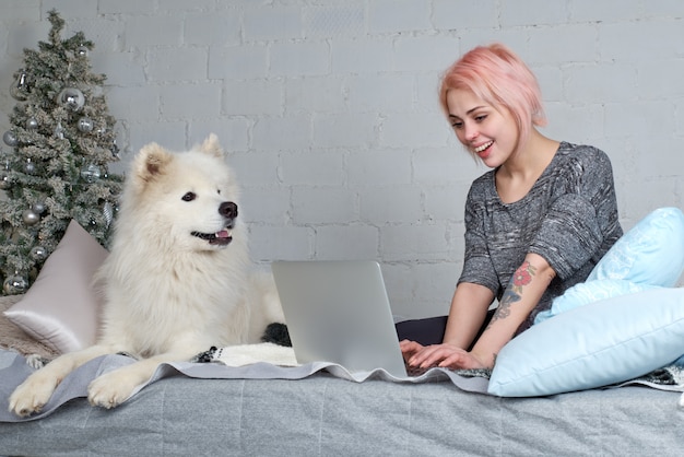 Jong mooi meisje met blond haar dat met laptop aan de bank met zijn grote witte hond werkt. Kerstboom en vreugdevolle uitdrukking.