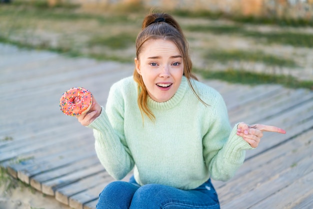 Jong mooi meisje met een donut in de buitenlucht verrast en wijzende vinger naar de zijkant