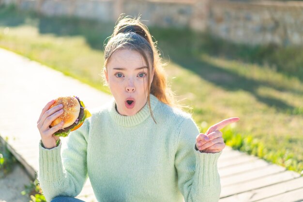 Jong mooi meisje met een hamburger in de open lucht verrast en wijzende kant