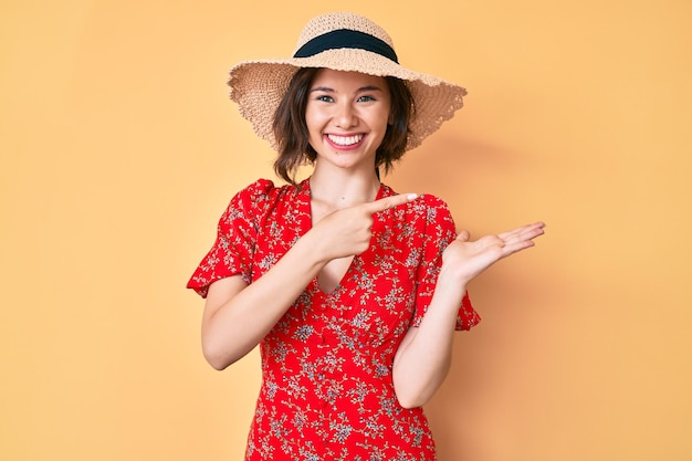 Jong mooi meisje met zomerhoed verbaasd en glimlachend naar de camera terwijl ze met de hand presenteert en met de vinger wijst.