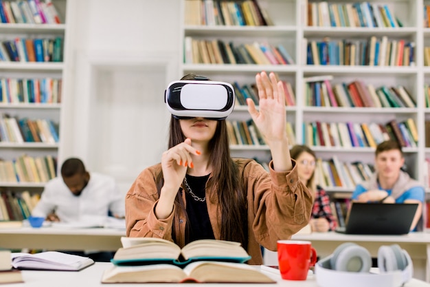 Jong mooi meisje, student, bibliotheekgebruiker, die VR-simulator gebruiken terwijl het doen van onderzoek naar bibliotheek. Vrouw die virtuele werkelijkheidsglazen dragen, die bij bureau met veel verschillende boeken zitten