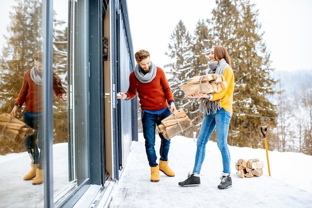 Jong mooi stel gekleed in kleurrijke truien die hun moderne huis binnenkomen met brandhout in de bergen tijdens de winter