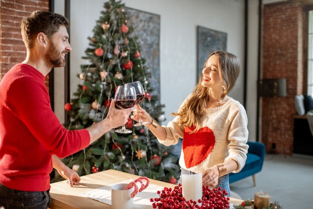 Jong mooi stel gekleed in truien die Nieuwjaar vieren en wijn drinken in het appartement met kerstboom
