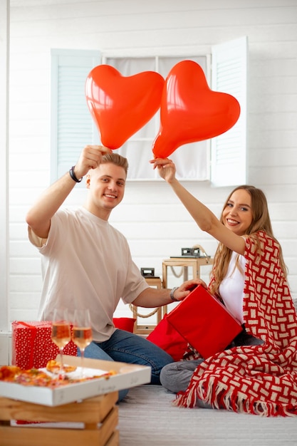 Jong mooi stel viert Valentijnsdag thuis, comfortabel zittend met deken, pizza en glazen wijn. Ze houden rode hartvormige ballonnen en geschenken vast. Liefde en geluk. Liefhebbers.