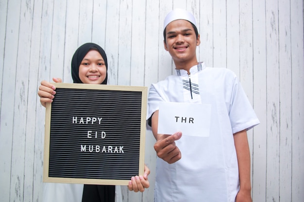 Jong moslimpaar met witte envelop genaamd THR Tunjangan Hari Raya en briefbord zegt Happy Eid Mubarak