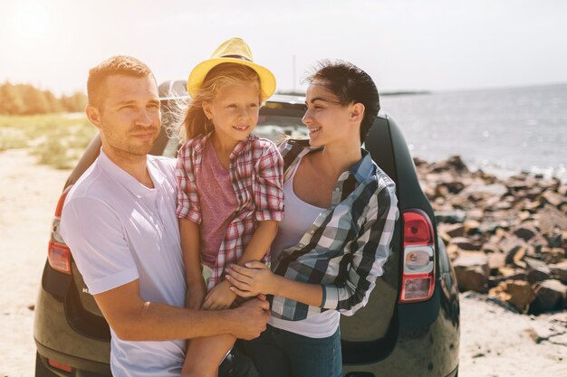 Jong paar dat zich dichtbij de geopende autolaars bevindt met koffers en zakken. Vader, moeder en dochter reizen door de zee of de oceaan of de rivier. Zomerrit met de auto