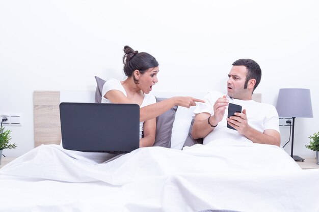 Jong paar die laptop en mobiele telefoon in bed thuis concept met behulp van