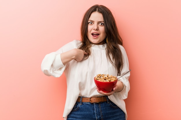 Jong plus size bochtige vrouw met een ontbijtgranen kom verrast wijzend op zichzelf, breed lachend.