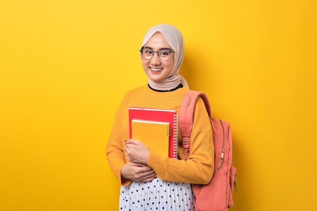 Jong positief Aziatisch studentenmeisje dat met het notitieboekje van de rugzakholding bij camera glimlacht die over gele achtergrond wordt geïsoleerd