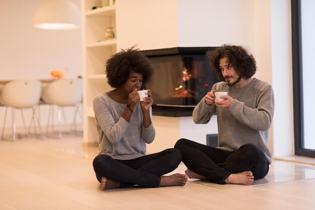 Jong romantisch multi-etnisch stel zittend op de vloer voor open haard thuis, elkaar aankijkend, pratend en koffie drinkend op herfstdag