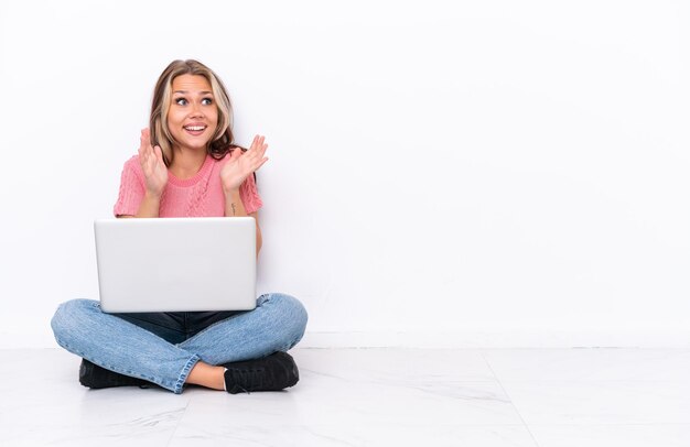 Jong Russisch meisje met een laptop zittend op de vloer geïsoleerd op een witte achtergrond met verrassing gezichtsuitdrukking