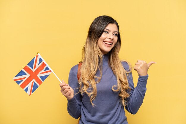 Jong Russisch meisje met een vlag van het Verenigd Koninkrijk geïsoleerd op een gele achtergrond, wijzend naar de zijkant om een product te presenteren