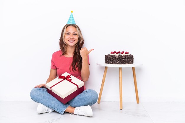 Jong Russisch meisje viert een verjaardag zittend op de vloer geïsoleerd op een witte achtergrond, wijzend naar de zijkant om een product te presenteren