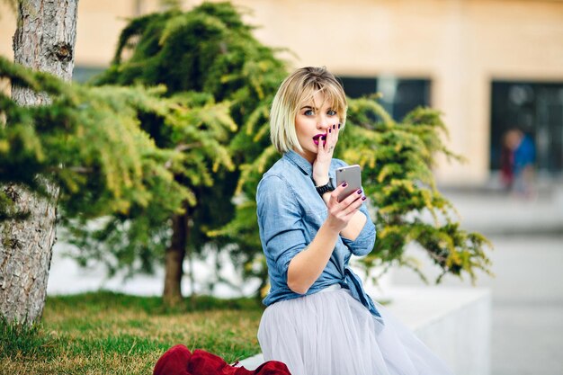 .Jong schattig blond meisje met kort haar en felroze lippen zittend in een park en het lezen van een bericht op haar smartphone en kijken geschokt gekleed in denimblauw shirt en grijze tule rok.