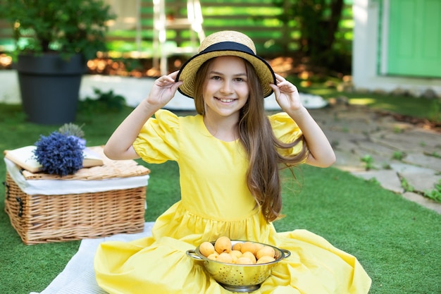 Jong schattig meisje houdt hoed met beide handen vast en zit met abrikozen in een vergiet bij picknick in de achtertuin