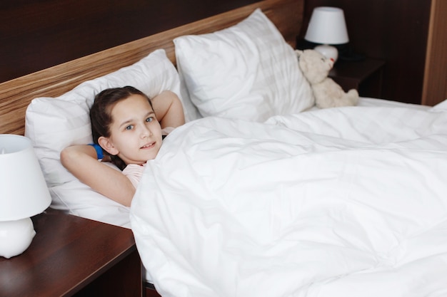 Jong schattig tienermeisje ligt in bed met witte kleren en kijkt naar de camera