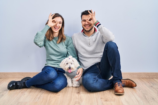 Jong spaans koppel zittend op de vloer met hond die ok gebaar doet met hand glimlachend, oog kijkend door vingers met blij gezicht.