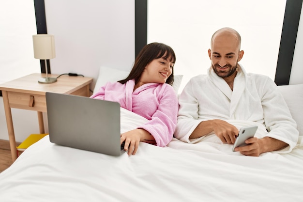 Jong spaans stel met behulp van laptop en smartphone zittend op het bed in de slaapkamer.