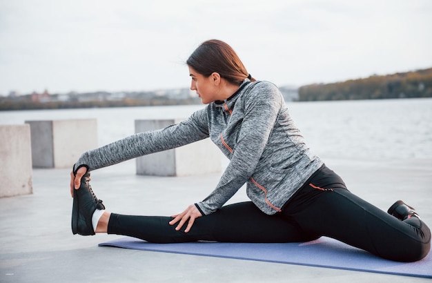 Foto jong sportief meisje doet yoga-oefeningen op fitnessmat buiten in de buurt van het meer overdag