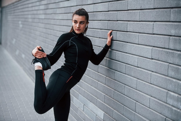 Jong sportief meisje in zwarte sportkleding buitenshuis die zich uitstrekt in de buurt van grijze muur