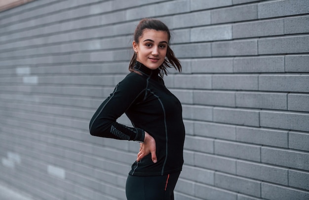 Jong sportief meisje in zwarte sportkleding die buiten in de buurt van grijze muur staat