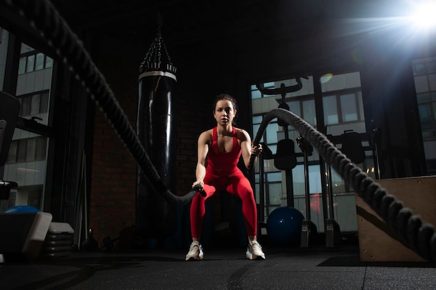 Jong sportmeisje in rode sportkleding traint met touwen in een zwarte donkere sportschool