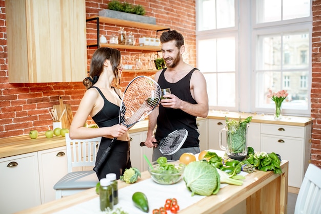 Jong sportpaar met een snack met vegetarisch eten drinken smoothie staande met rackets op de keuken thuis