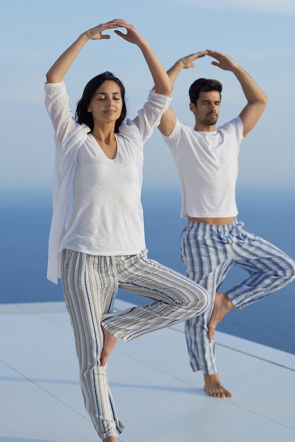 jong stel dat yoga beoefent bij zonsondergang in een modern huisterras met oceaan en zonsondergang op de achtergrond
