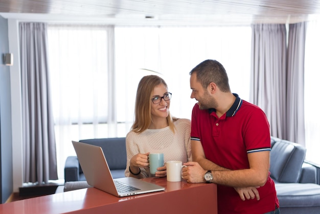 Jong stel drinkt koffie en gebruikt laptopcomputer in luxe huis samen, kijkend naar het scherm, glimlachend.