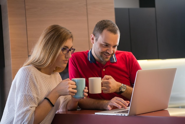 Jong stel drinkt koffie en gebruikt laptopcomputer in luxe huis samen, kijkend naar het scherm, glimlachend.