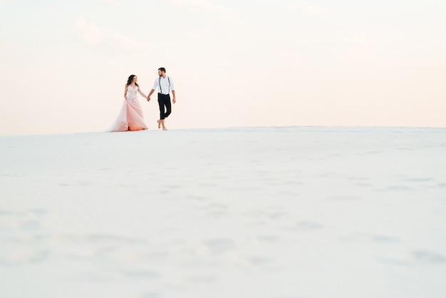 Jong stel een man in zwarte broek en een meisje in een roze jurk lopen langs het witte zand van de woestijn